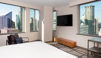 A modern bedroom with large windows offering a city view, a wall-mounted TV, and minimal furnishings including a chair, table, and bench.