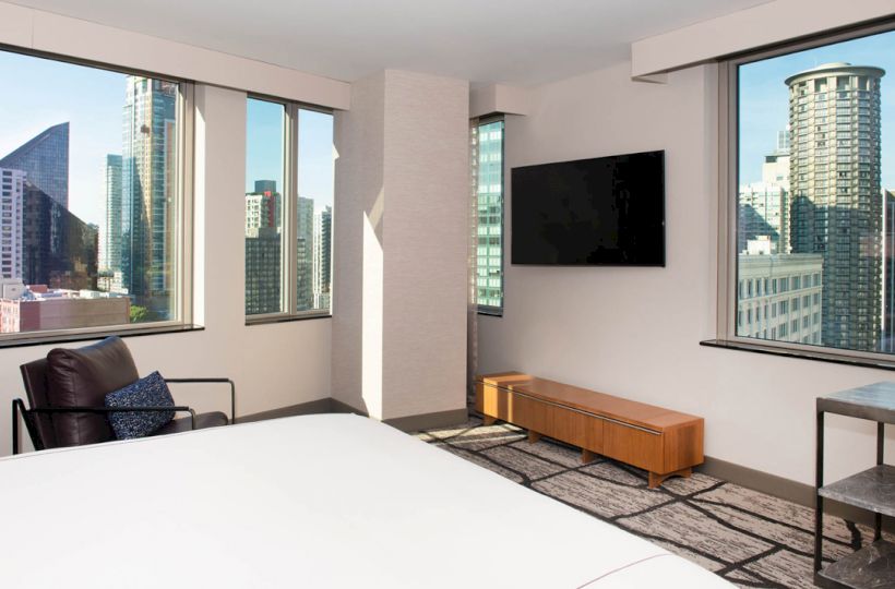 A modern bedroom with large windows offering a city view, a wall-mounted TV, and minimal furnishings including a chair, table, and bench.