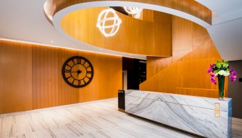 A modern lobby features a marble reception desk, a large decorative clock on a wooden wall, and stylish lighting fixtures above.