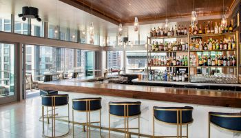 This image shows a modern bar with a wooden counter, bar stools, a variety of bottled beverages on shelves, and floor-to-ceiling windows.