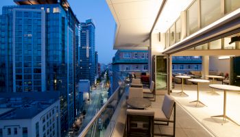 A modern cityscape at dusk featuring tall buildings and a well-lit balcony with seating and tables overlooking a busy street below.