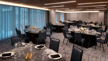 The image shows an elegantly set banquet hall with round tables, black chairs, floral centerpieces, and blue curtains, lit by modern ceiling lights.