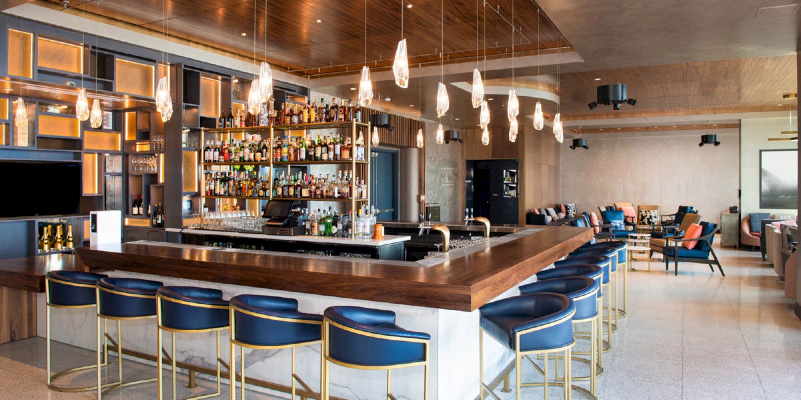 A stylish bar with a wooden counter, blue and gold bar stools, pendant lights, and a well-stocked back bar. The seating area is in the background.