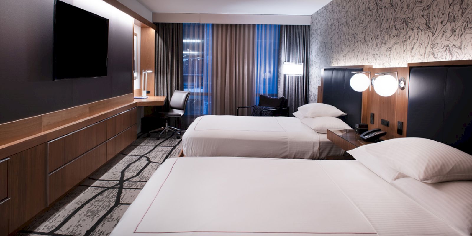 A modern hotel room with two beds, a wall-mounted TV, a desk with a chair, stylish wallpaper, and soft lighting over the beds and desk.