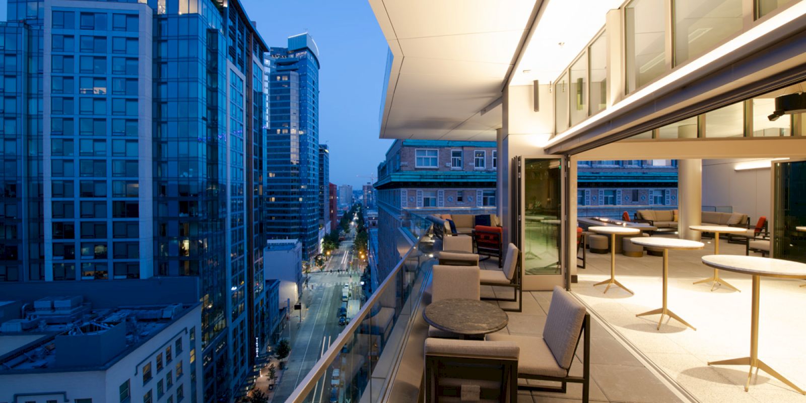 A modern cityscape with high-rise buildings at dusk, featuring a sleek balcony with tables and chairs overlooking the street below.