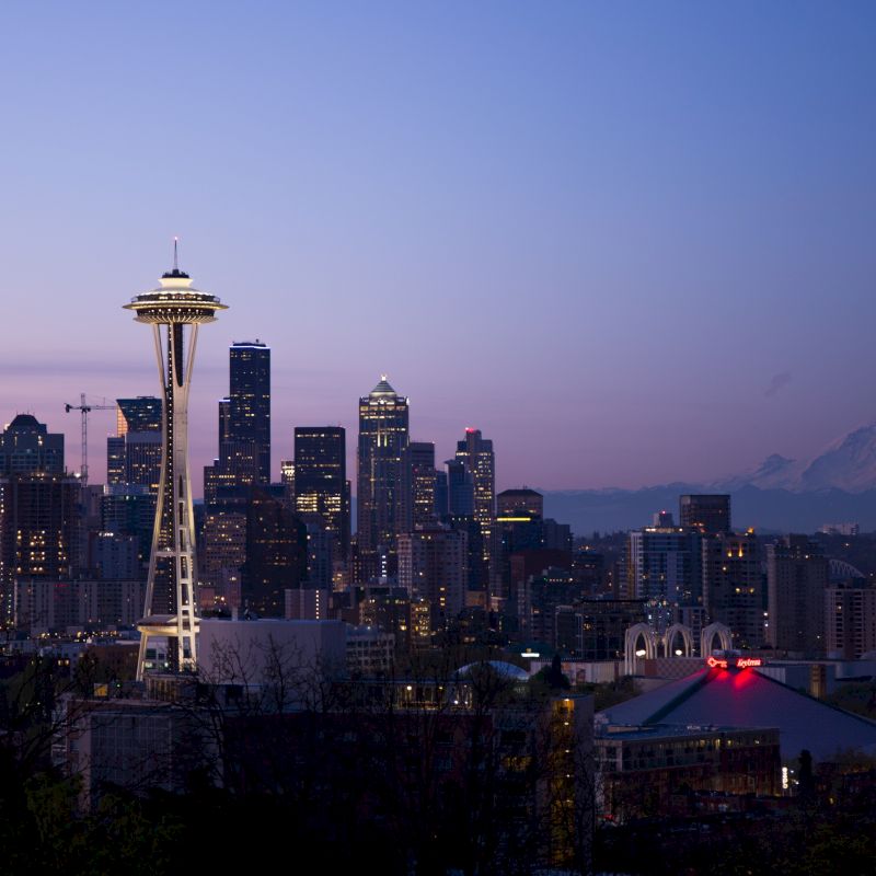 The image shows a city skyline at dusk, featuring the Space Needle prominently, with tall buildings and Mount Rainier visible in the background.