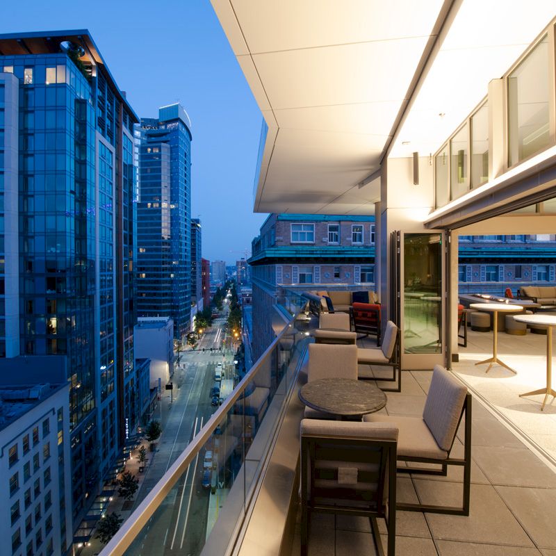A cityscape view from a modern, urban balcony with seating and tables, high-rise buildings illuminated as night falls.