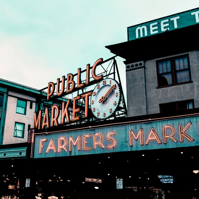 A neon sign reading "Public Market" and "Farmers Market" with a clock is displayed on a historic building, likely representing Pike Place Market.