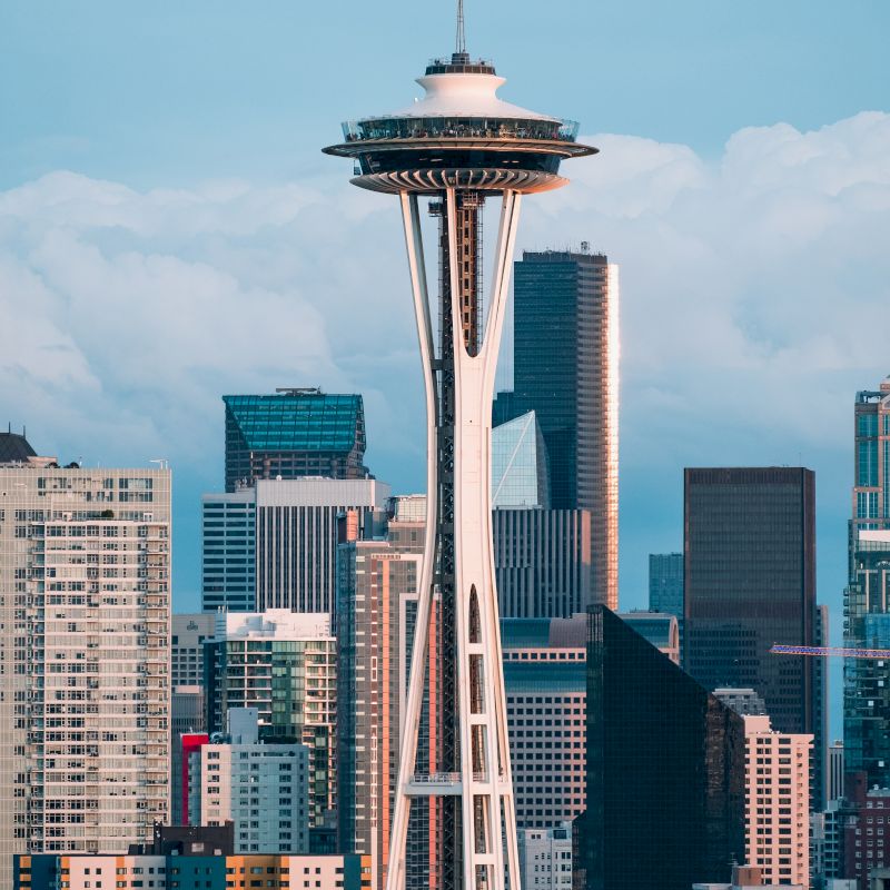 The image showcases the Space Needle, an iconic observation tower, with the Seattle skyline and various skyscrapers in the background, under a partly cloudy sky.