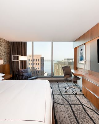 A modern hotel room with a large bed, a desk, a chair, and a wide window offering a view of buildings. The room has a wall-mounted TV.