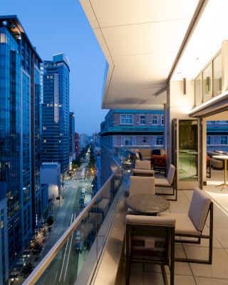 A modern cityscape featuring high-rise buildings at dusk, with a sleek, well-lit rooftop terrace equipped with tables and chairs for seating.