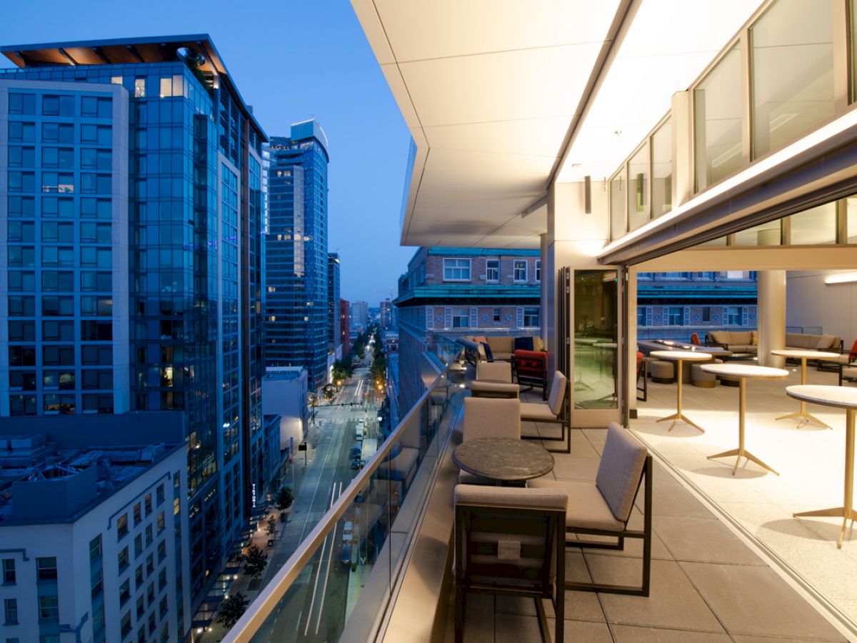 A modern high-rise city view from a balcony at dusk, featuring outdoor seating and tables. Tall buildings with glass windows line the street below.