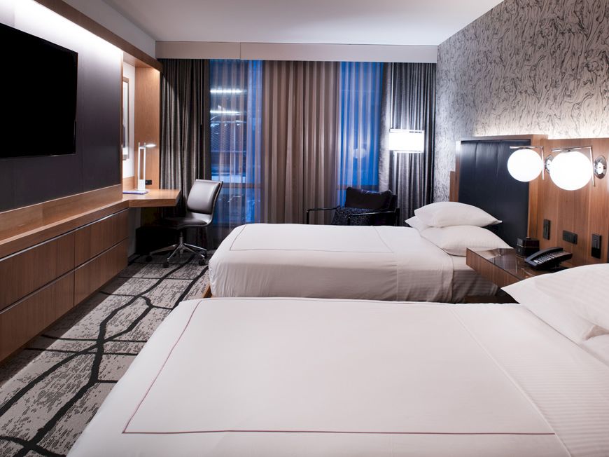 A modern hotel room with two neatly-made beds, a wall-mounted TV, a desk by the window, and contemporary lighting and decor.