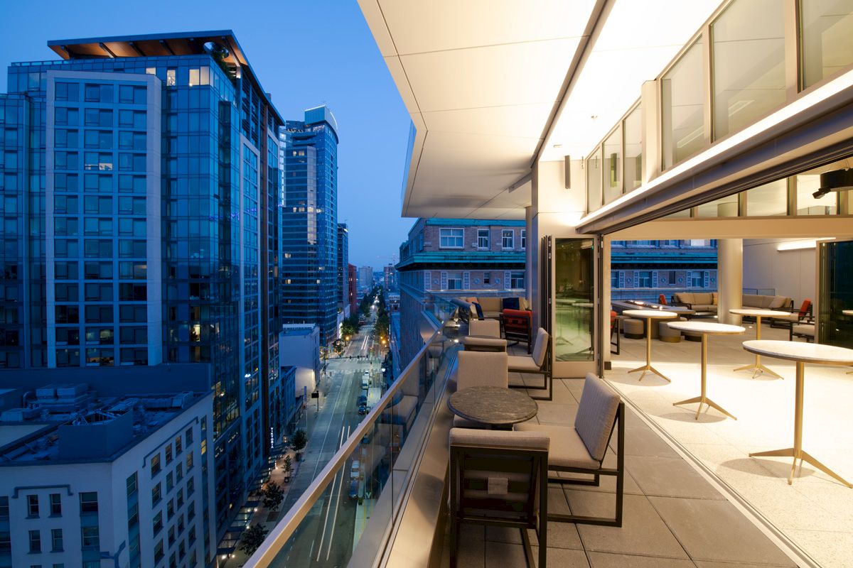A modern urban balcony overlooks a city street with high-rise buildings at dusk, featuring outdoor seating and tables.