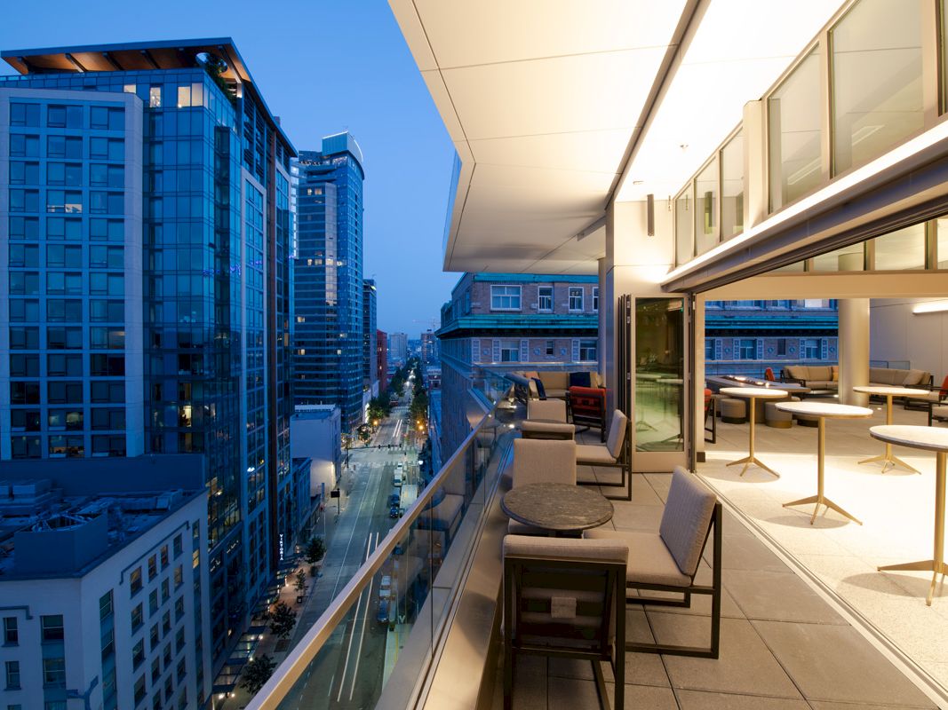 A modern urban balcony overlooks a city street with high-rise buildings at dusk, featuring outdoor seating and tables.