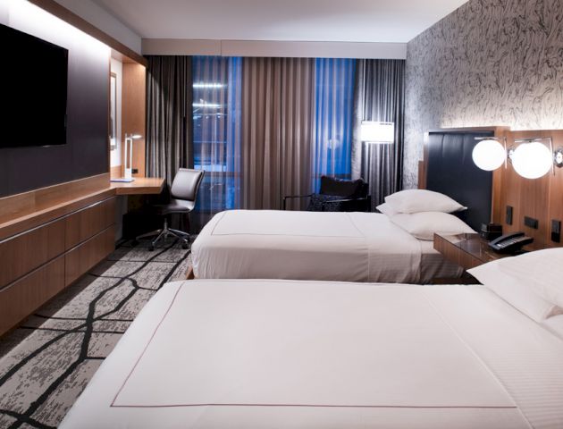 The image shows a modern hotel room with two neatly made beds, a wall-mounted TV, a desk with a chair, and contemporary decor.