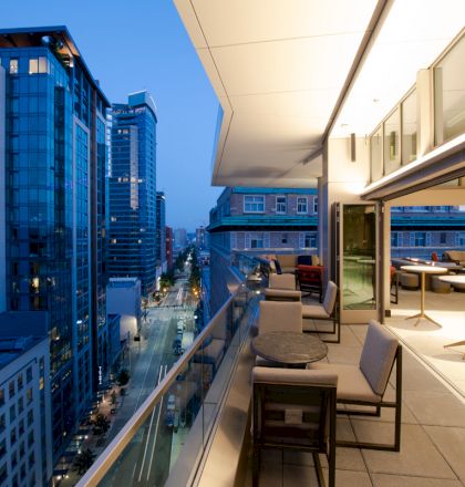 A modern cityscape view from a high balcony, featuring outdoor seating and buildings with glass facades during dusk.