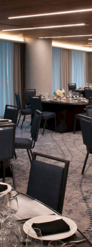 A banquet room setup with round tables covered in black tablecloths, set with tableware, glassware, napkins, and floral centerpieces, under warm lighting.