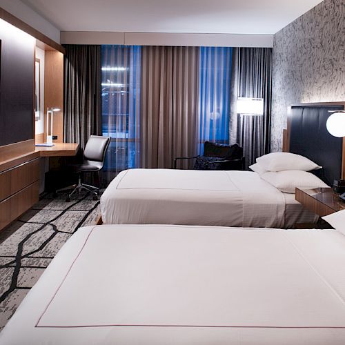 A modern hotel room with two double beds, a wall-mounted TV, a work desk with a chair, and stylish lighting, providing a comfortable stay.