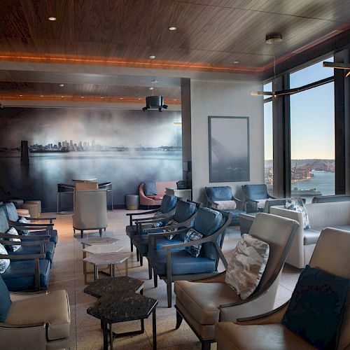 This image shows a modern lounge area with plush seating, a cityscape mural, and large windows providing a scenic view of a waterfront and Ferris wheel.