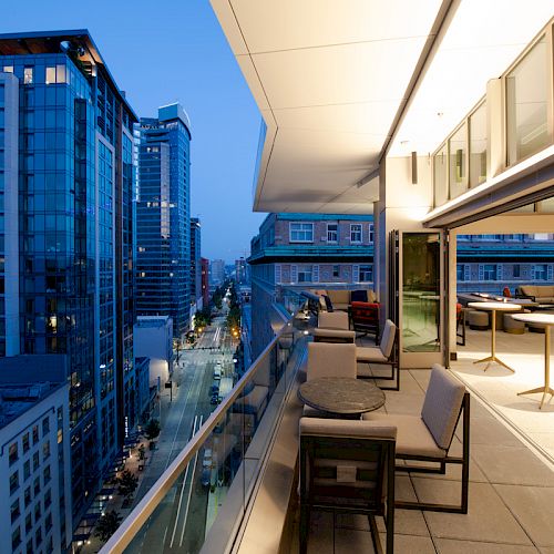 A modern cityscape view from a balcony at dusk, featuring tall buildings, street lights, and a sleek outdoor seating area with tables and chairs.