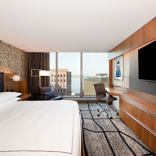 A modern hotel room features a large bed, a desk with a chair, a flat-screen TV, and a window with a city view.