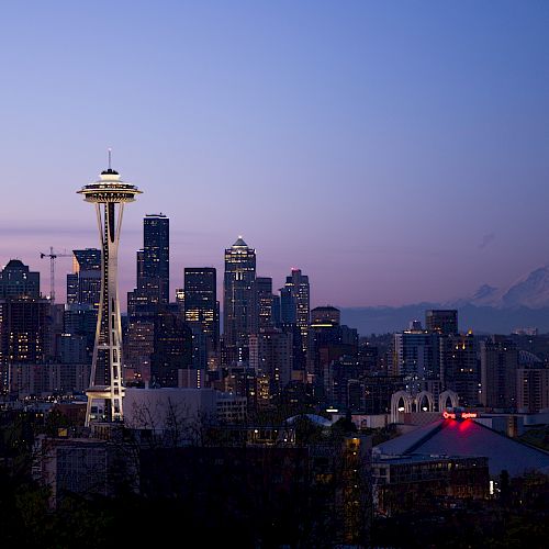 A city skyline at dusk, featuring a prominent tower and skyscrapers, with a distant mountain in the background under a twilight sky.
