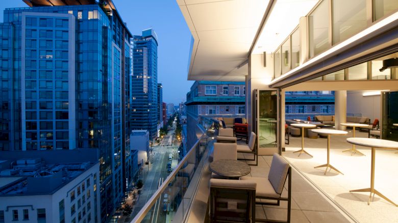 A modern cityscape with tall buildings and a well-lit outdoor patio area featuring tables and chairs, overlooking a city street at twilight.