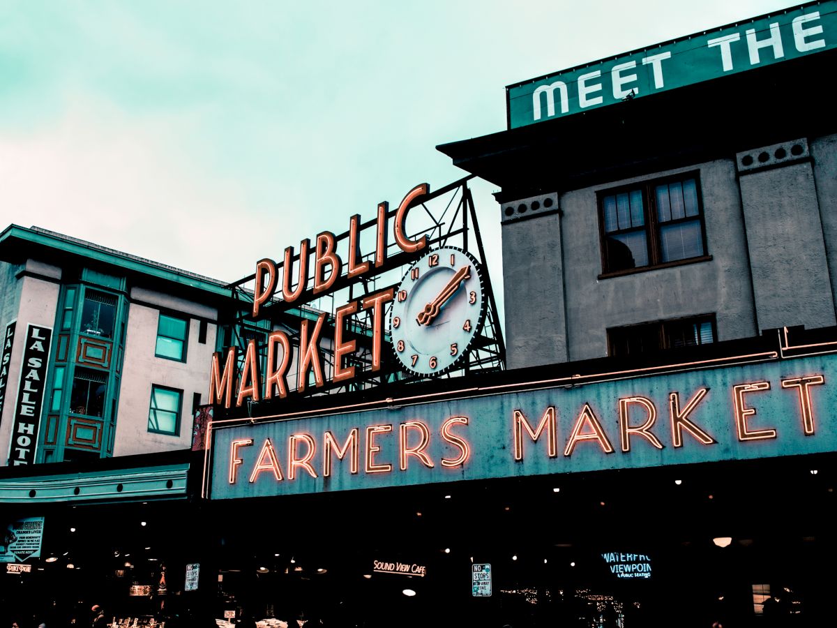 The image shows a public market with a neon sign that reads "Public Market" and "Farmers Market." There's a clock in the center and nearby buildings.