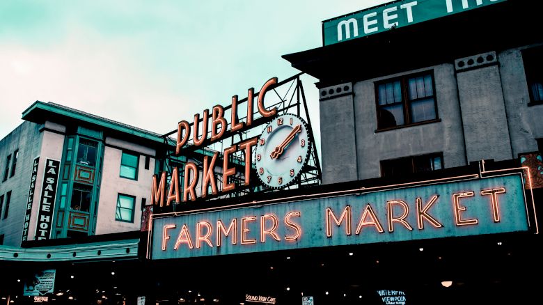 The image shows a public market with a neon sign that reads "Public Market" and "Farmers Market." There's a clock in the center and nearby buildings.
