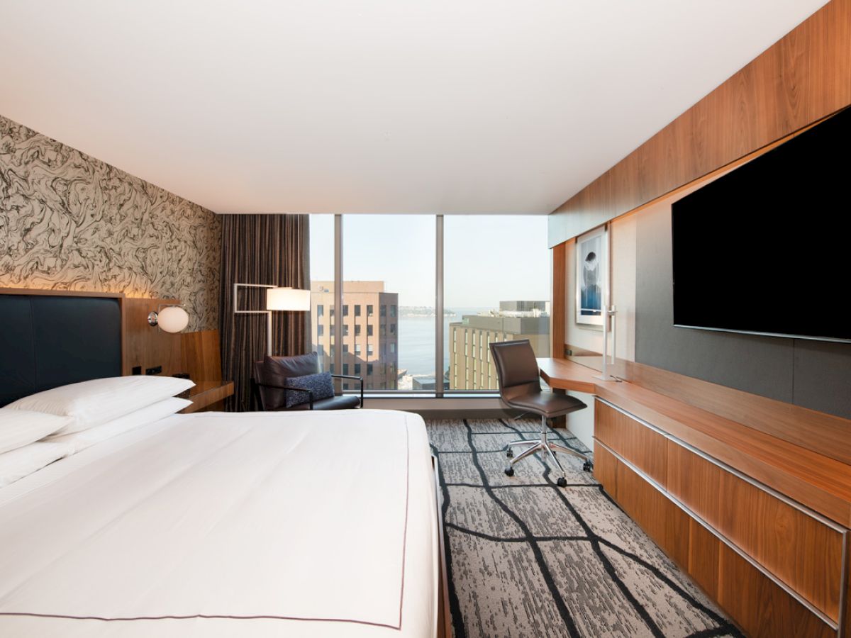 A modern hotel room features a large bed, a desk with a chair, a wall-mounted TV, and a window with a view of city buildings and water.