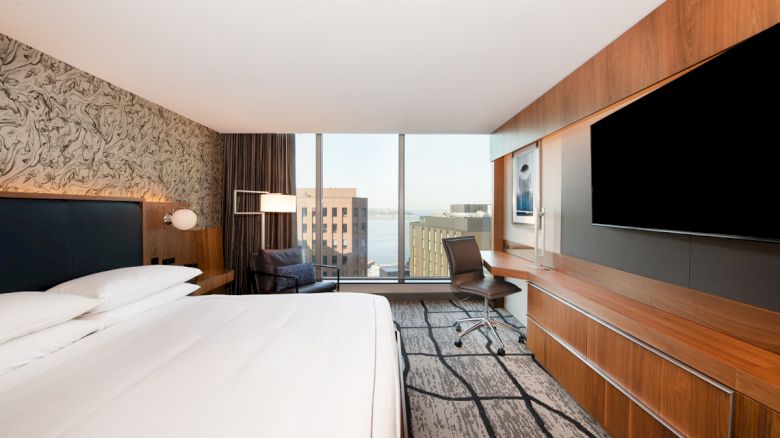 A modern hotel room features a large bed, a desk with a chair, a wall-mounted TV, and a window with a view of city buildings and water.