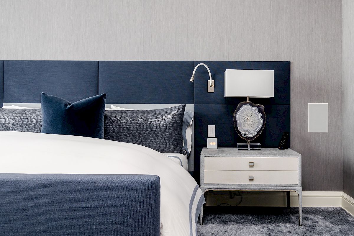 This image shows a modern bedroom with a blue upholstered bed, gray and white bedding, a bedside table with a lamp, and a decorative clock.