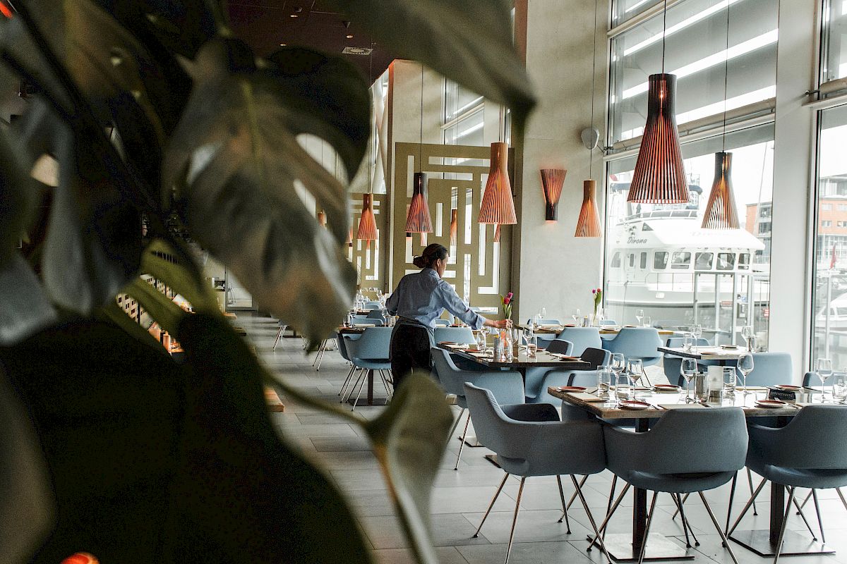 A person is setting the table in a modern restaurant with stylish pendant lights and large windows, with some plants in the foreground.