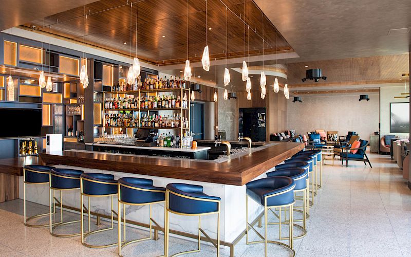 The image shows a modern bar with stylish lighting, blue bar stools, a well-stocked shelf of liquor bottles, and a cozy seating area in the background.