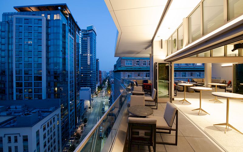 A modern cityscape view from a high-rise balcony at dusk, with comfortable seating and tables, overlooking illuminated buildings and streets.