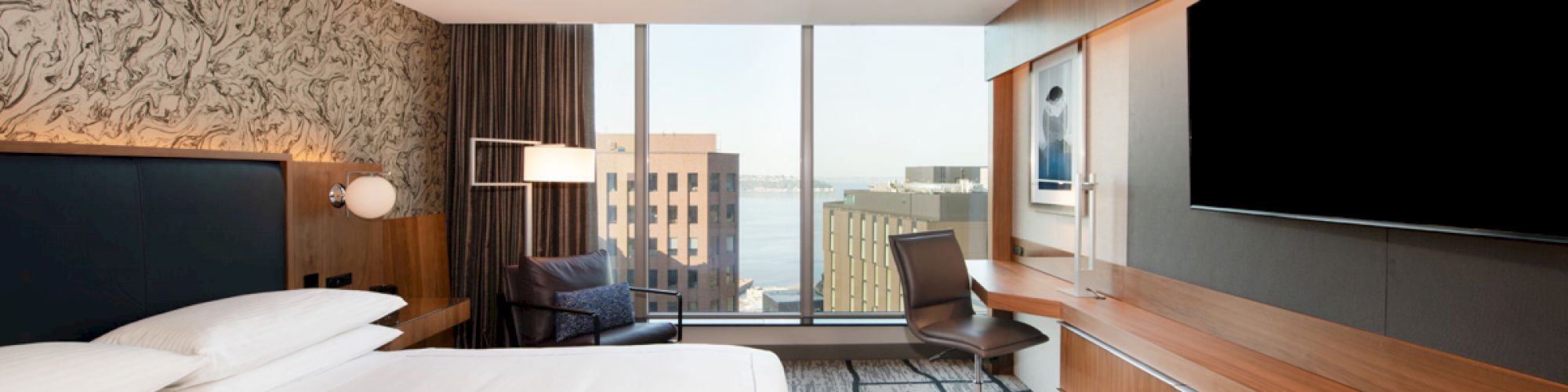 A modern hotel room with a large bed, desk, chair, and a flat-screen TV; a city view through large windows can be seen.